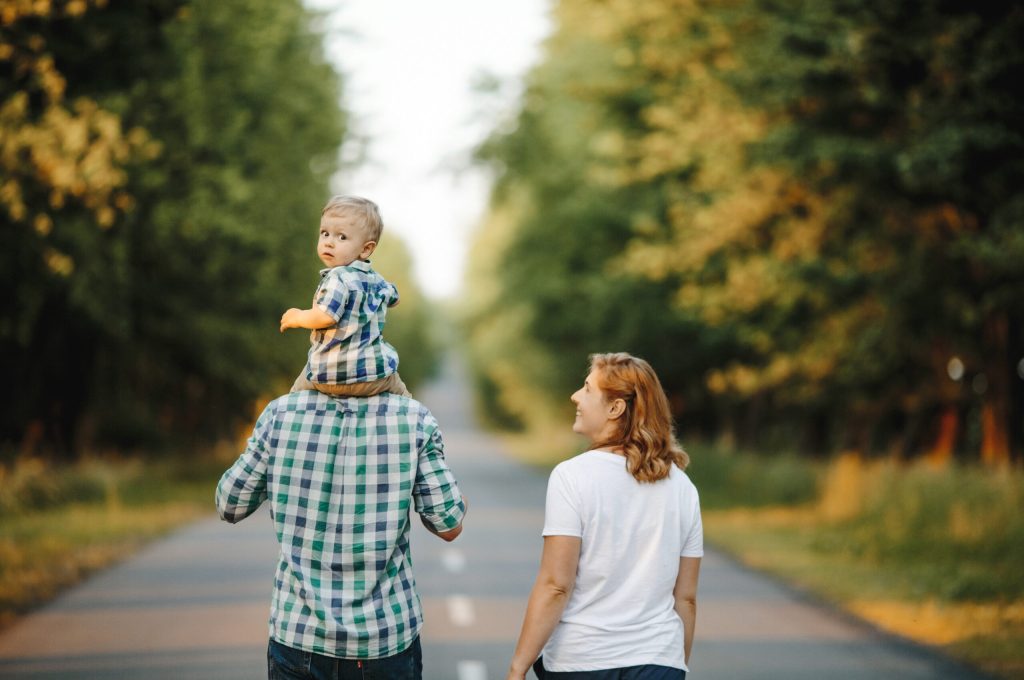 Father holds a son on their shoulders, a mother is nearby, they are walking, a little boy turns around